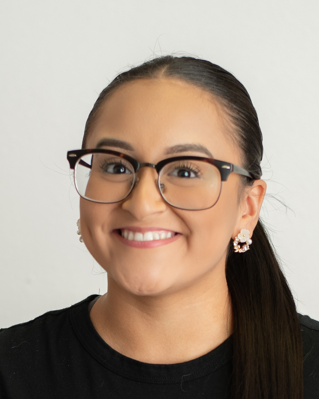 A woman with long dark hair tied back, wearing glasses, earrings, and a black shirt, smiling against a plain background.