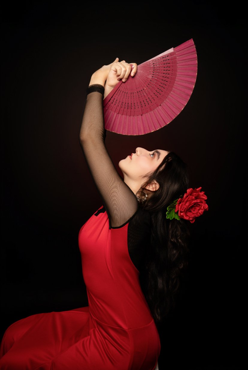 A woman dressed in a red outfit with black mesh sleeves, holding a pink fan above her head, against a black background. She has a large red flower in her hair and is looking up at the fan.