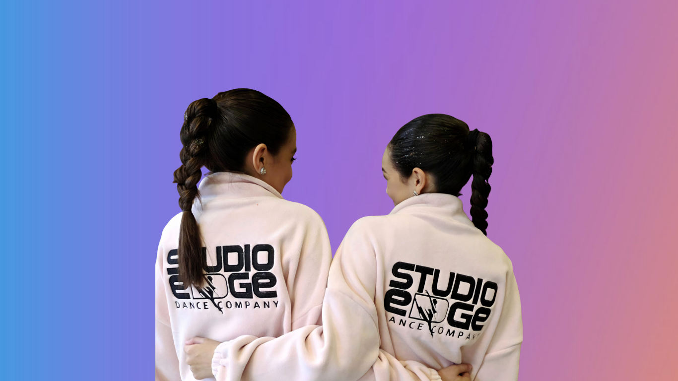 Two women with braided hair wearing cream-colored sweatshirts with 'Studio Edge Dance Company' logo on back, facing away from the camera against a gradient background.