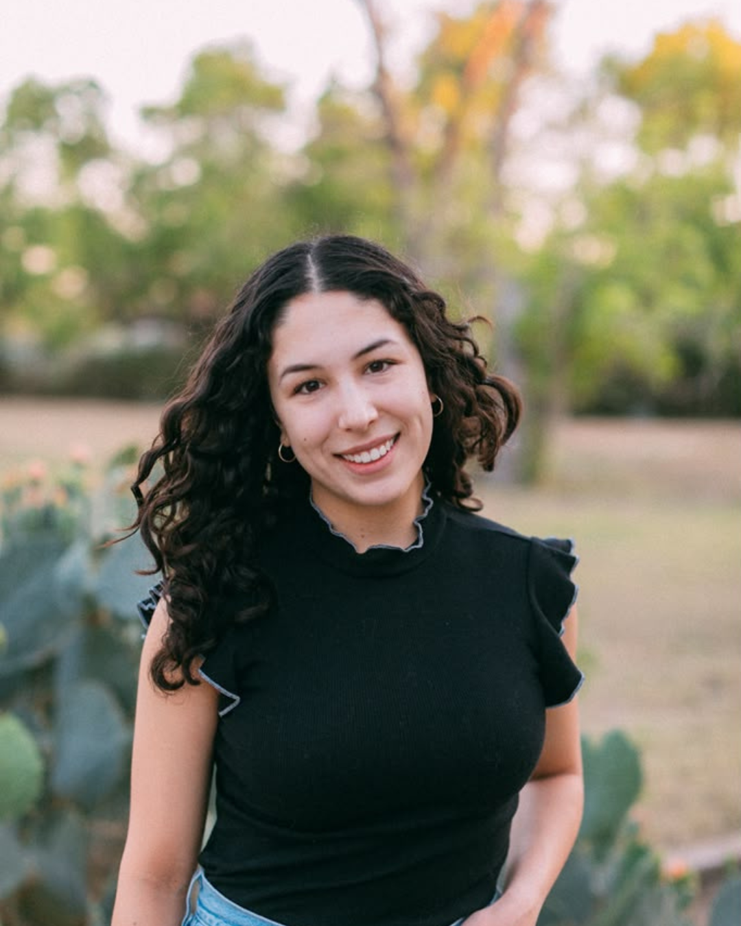 A young woman with dark curly hair, wearing a black sleeveless top with ruffled sleeves, smiling outdoors in a park or garden area during fall with trees and greenery in the background.