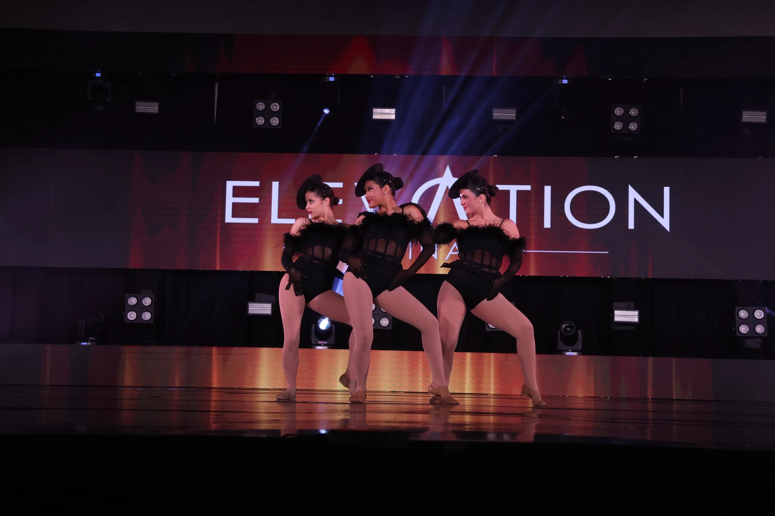 Four women performing a dance routine on stage with a large digital screen in the background displaying the word 'ELECTATION'. The women are dressed in black costumes with sheer and feather details, striking a pose with one leg raised.