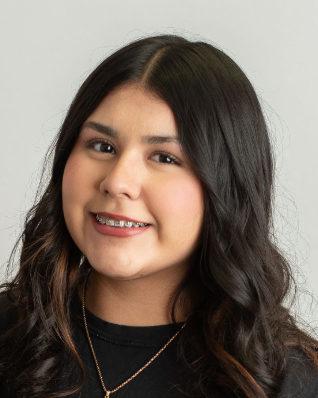 A young woman with long dark hair, smiling, wearing braces, a black top, and a gold necklace, posed against a plain background.