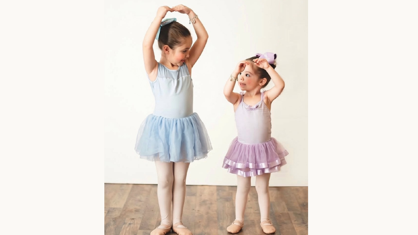 Two young girls in ballet tutus, one in blue and the other in pink, are playing and smiling at each other, touching their bows on their heads.