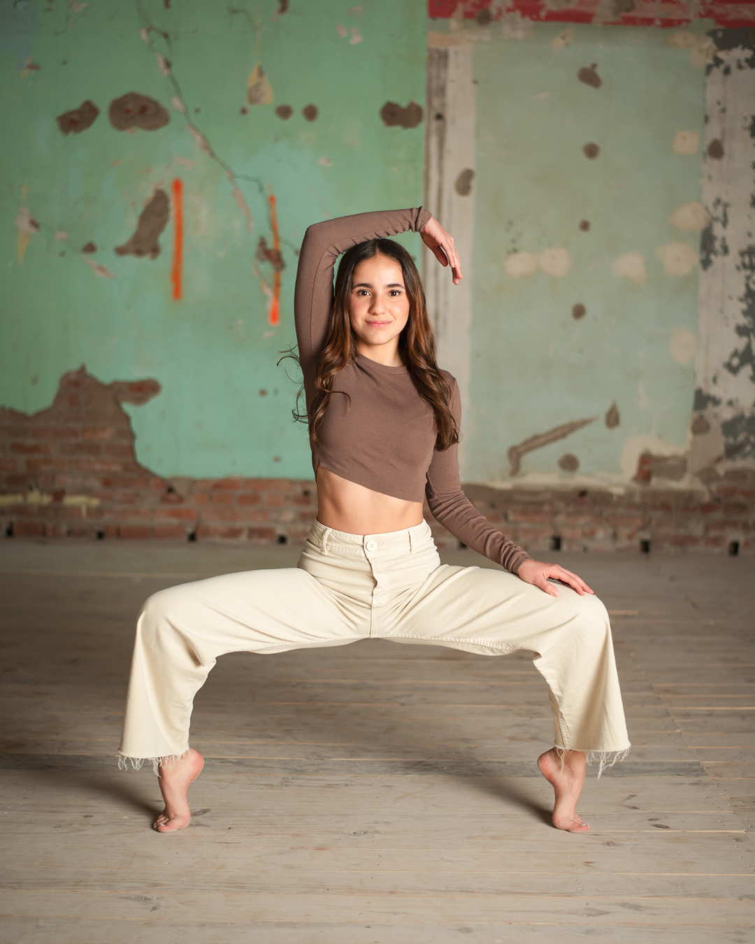 A young woman practicing yoga in an industrial-style room with a distressed green wall and wooden floor, standing in a wide-legged stance with one arm raised overhead.