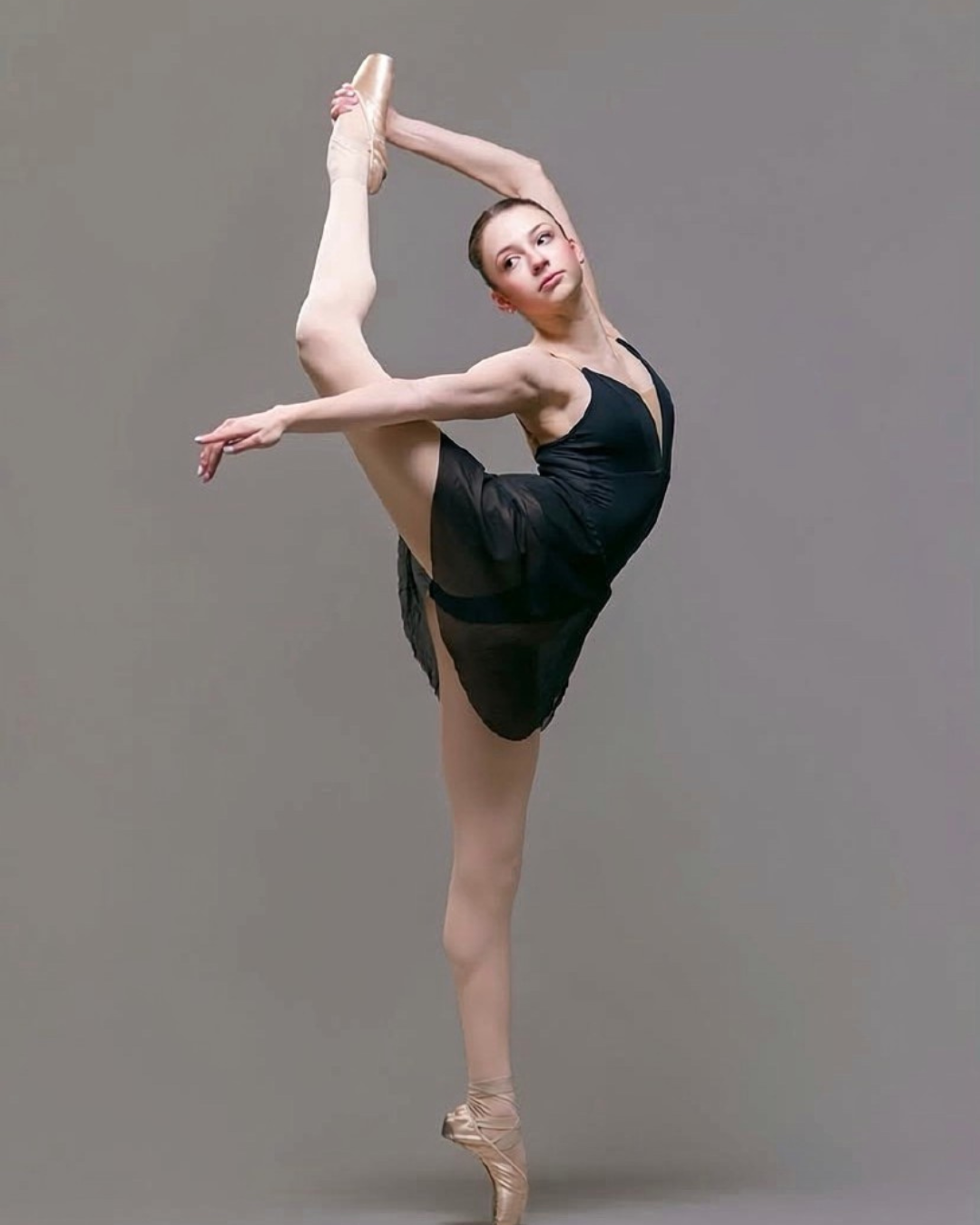 Ballet dancer in a black dress executing an advanced pose on pointe against a gray background.
