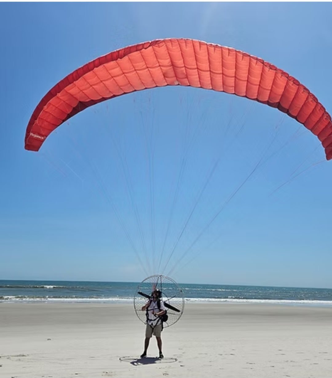 Person standing on beach preparing to fly a red parachute with a propeller backpack, blue sky and ocean in the background.