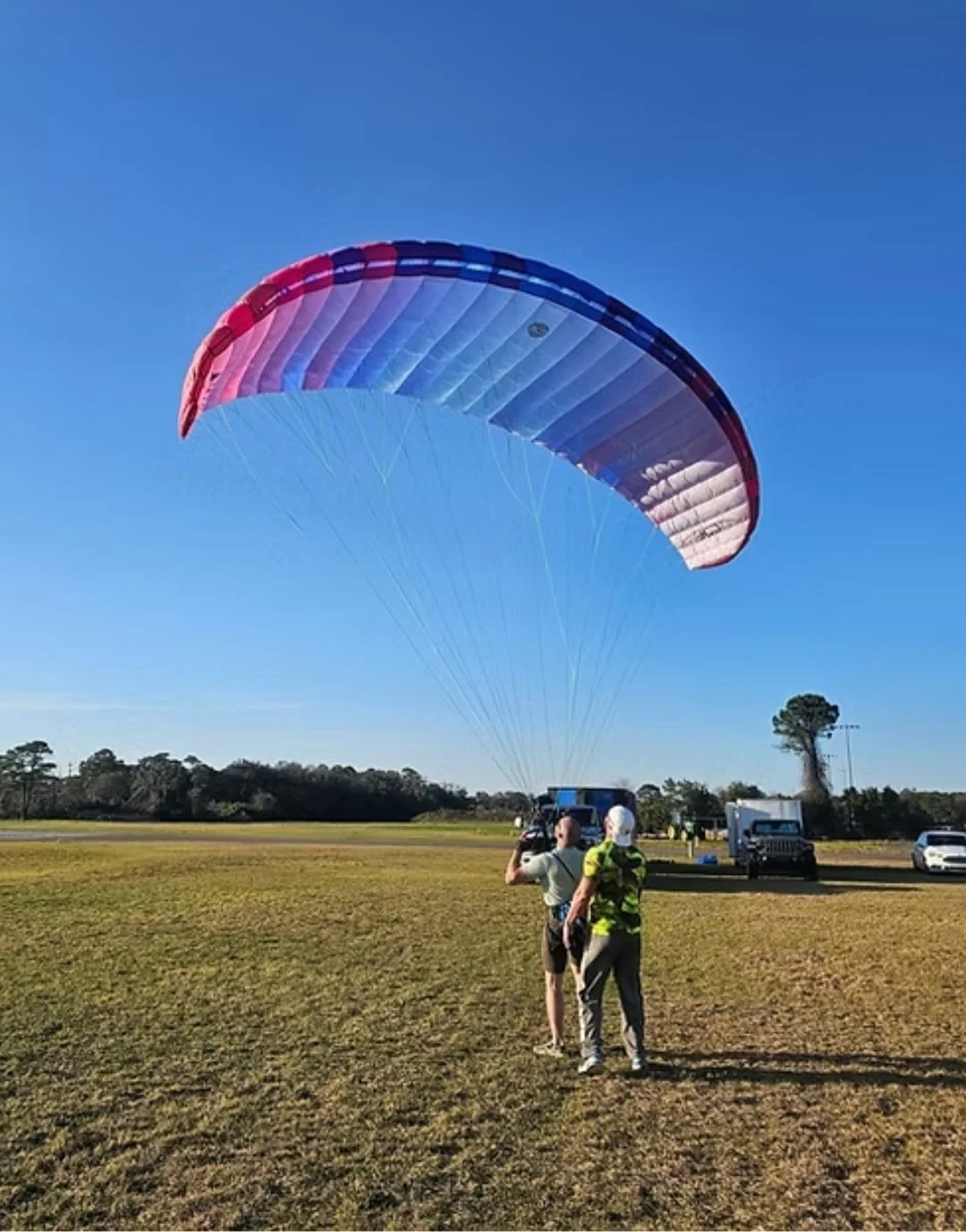 A person is preparing to paraglide on a large grassy field under a clear blue sky, with a support crew nearby and vehicles in the background.