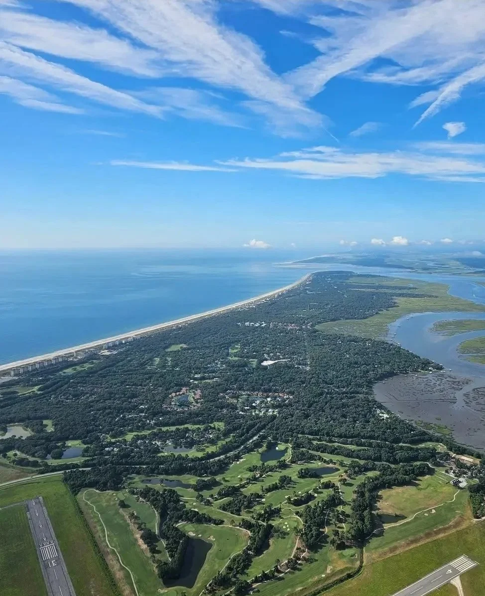 Aerial view of a coastal landscape showing a beach, ocean, and numerous green parks and waterways, with a small airport runway at the bottom left.