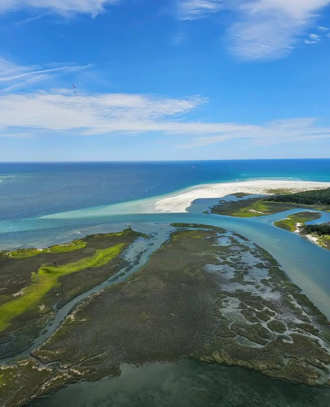 Aerial view of a coastal landscape showing wetlands, sandy beach, ocean, and sky with some clouds.