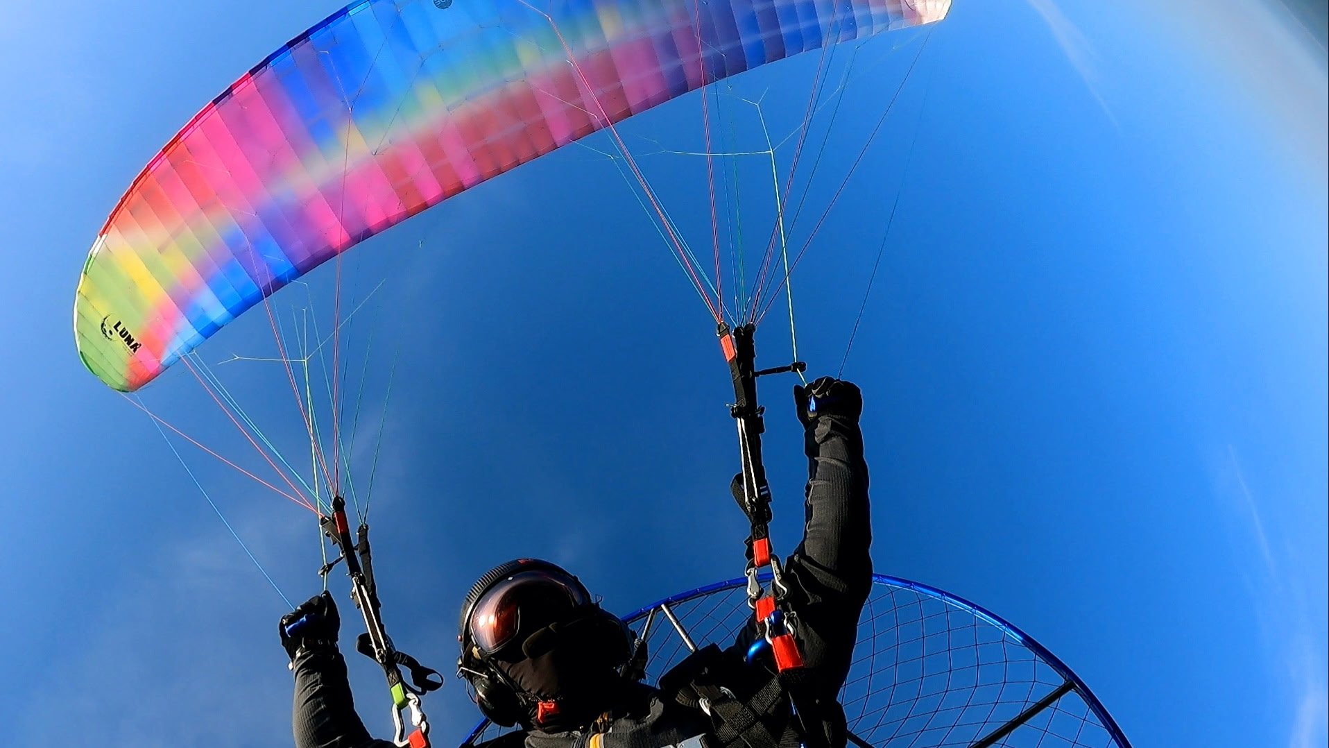 Person paragliding with a vibrant rainbow-colored wing against a clear blue sky.