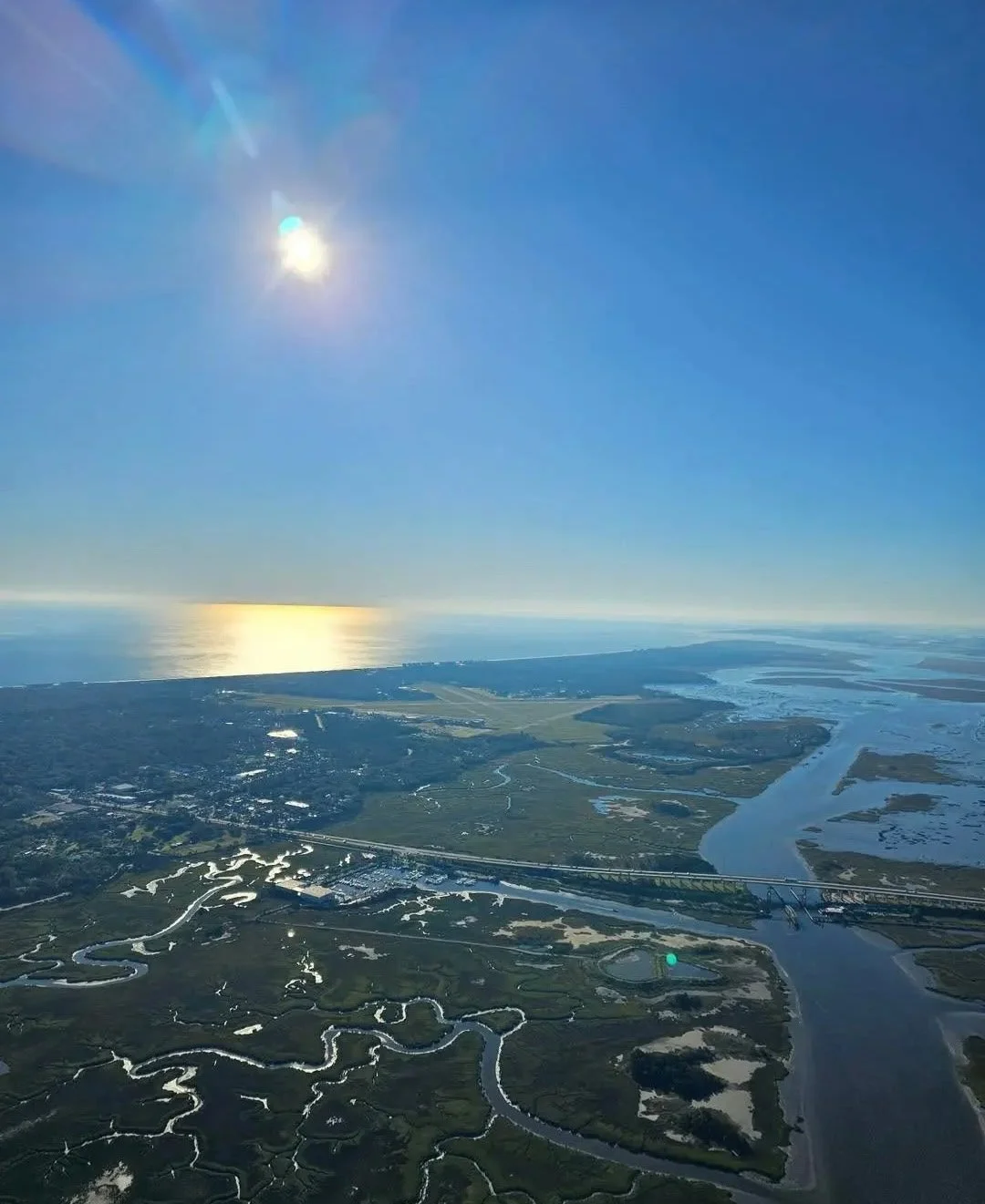 Aerial view of a coastal wetland landscape with winding waterways, a bridge, and the ocean in the distance under a clear blue sky with the sun shining.