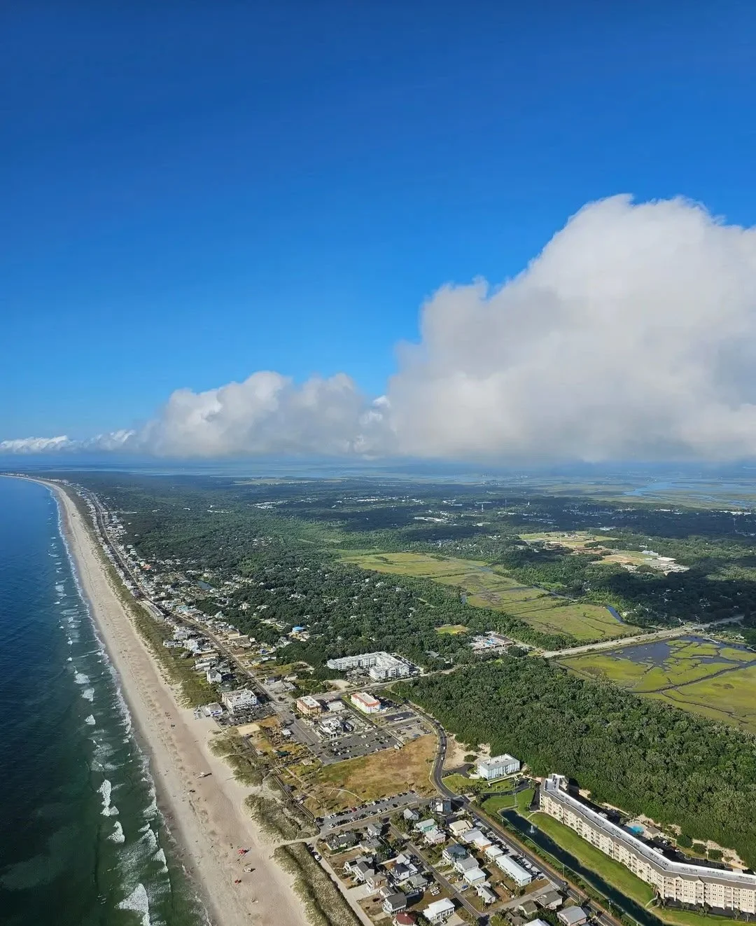 Aerial view of a coastal area with a sandy beach, ocean waves, and a town with buildings, surrounded by green fields and forests under a partly cloudy blue sky.