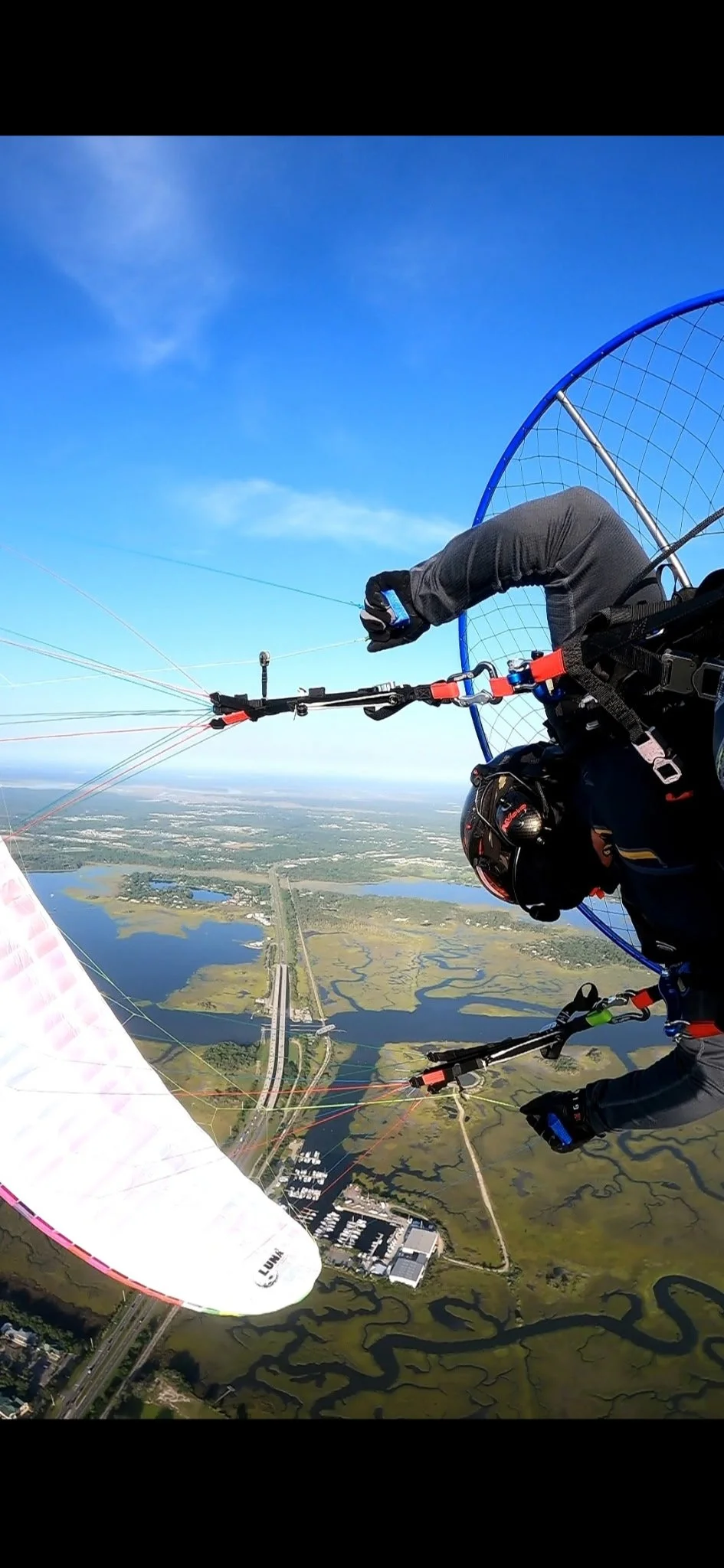 A person gliding through the sky while paragliding over a landscape with water bodies, roads, and green fields on a bright, sunny day.