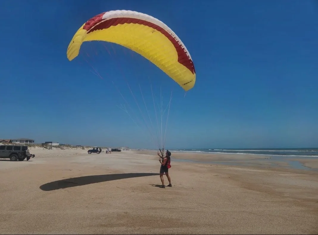 Person kiting a yellow and red paramotor wing on a sandy beach with parked cars and a clear blue sky.