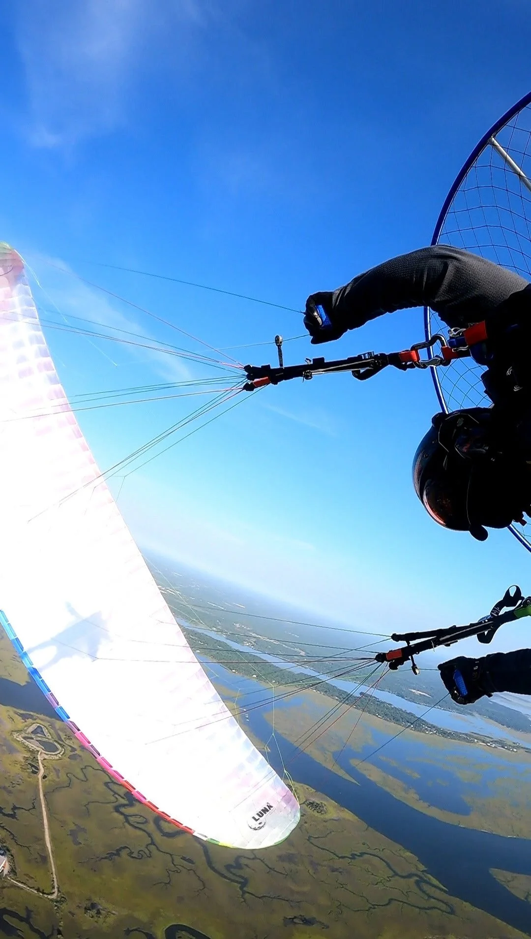 A person paragliding over a landscape with water bodies and fields, mid-air, with a colorful paraglider wing visible.
