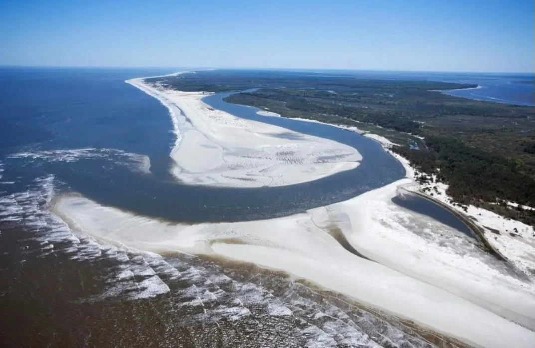 Aerial view of a winding river along a snow-covered coastline with open water and land visible in the background.