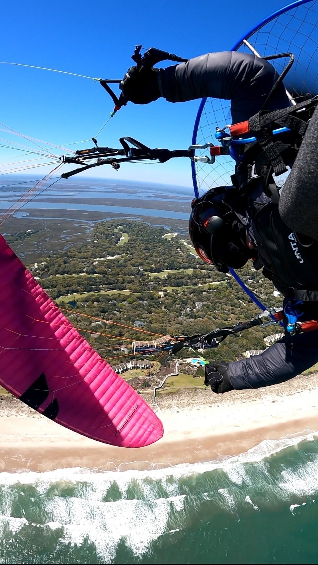 Paraglider flying upside down high above a beach with waves crashing below, holding a pink wing, with a view of green fields and water bodies in the distance, and a clear blue sky.