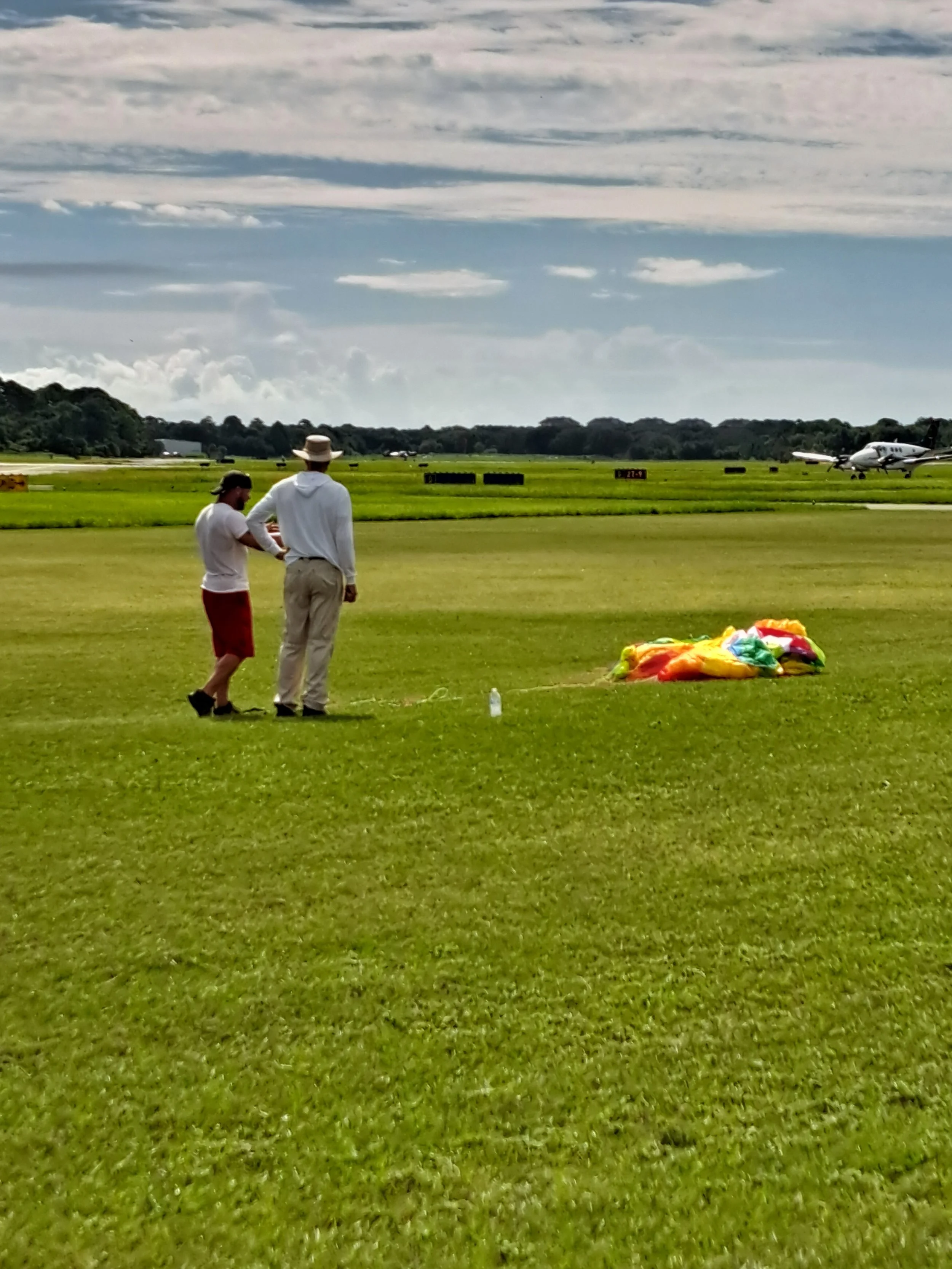 Two people standing on a grassy field discussing a paramotor wing laid out on the grass in front of them with an aircraft and airport signage in the background, under a cloudy sky.
