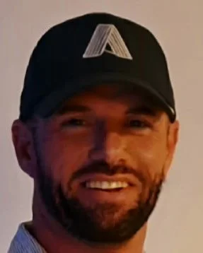 A man wearing a black baseball cap with a white geometric logo, smiling with a beard.