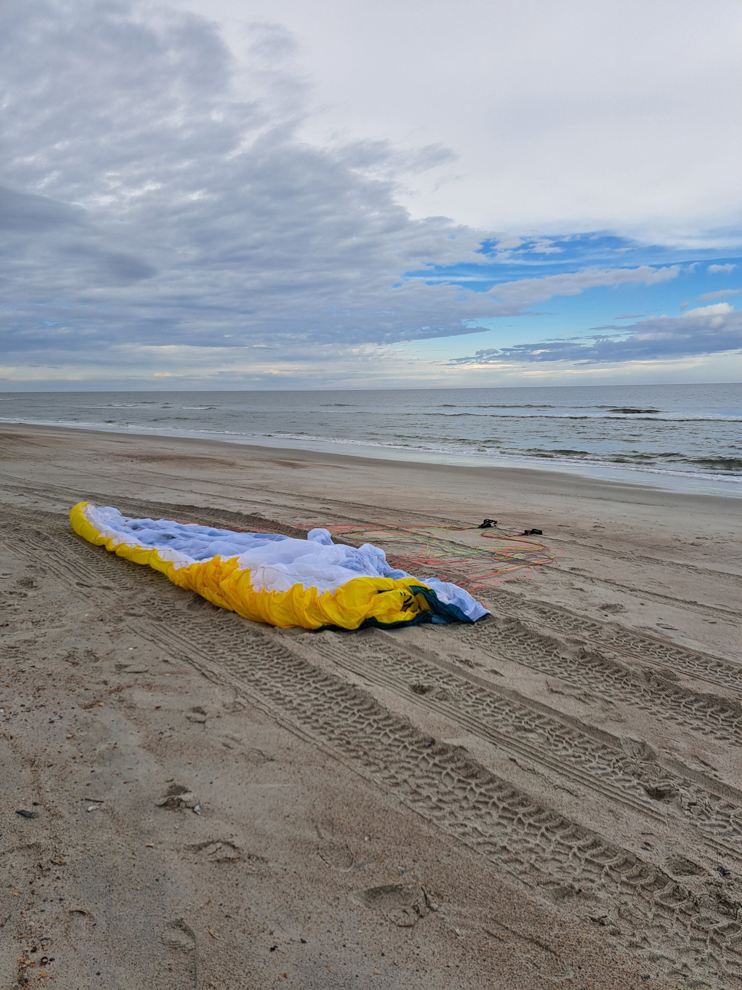 A deflated yellow and white parachute lies on the sandy beach near the shoreline, with colorful parasailing cords attached. The ocean waves gently reach the shore under a cloudy sky with some patches of blue.