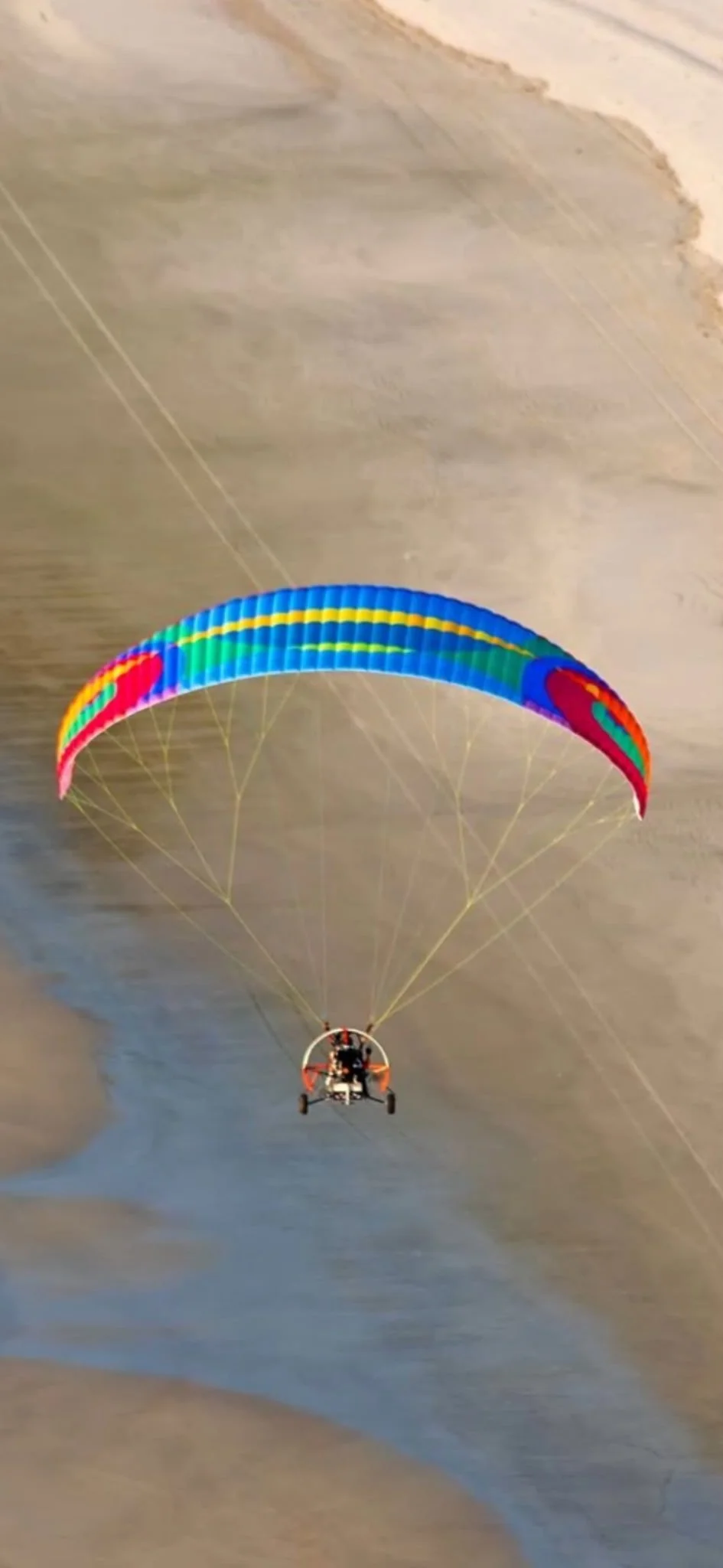 A person flying a colorful, rainbow-patterned parachute with a motorized trike against a cloudy sky.