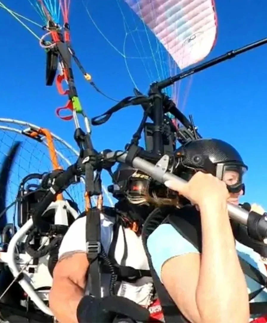 A person wearing a helmet and sunglasses preparing a powered harness for parachuting or paragliding against a clear blue sky.