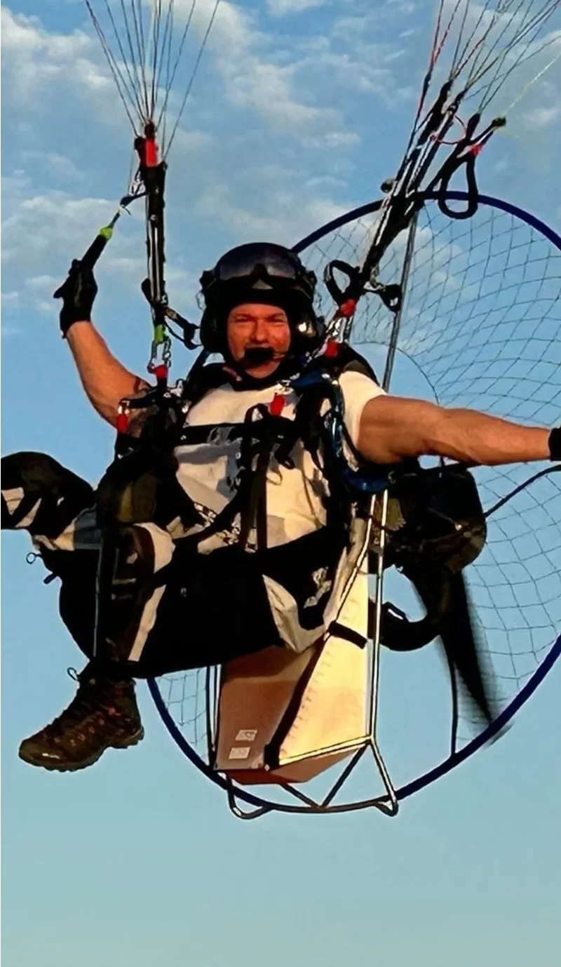 Person in a powered parachute flying in the sky during daytime, wearing a helmet and harness, with clouds in the background.
