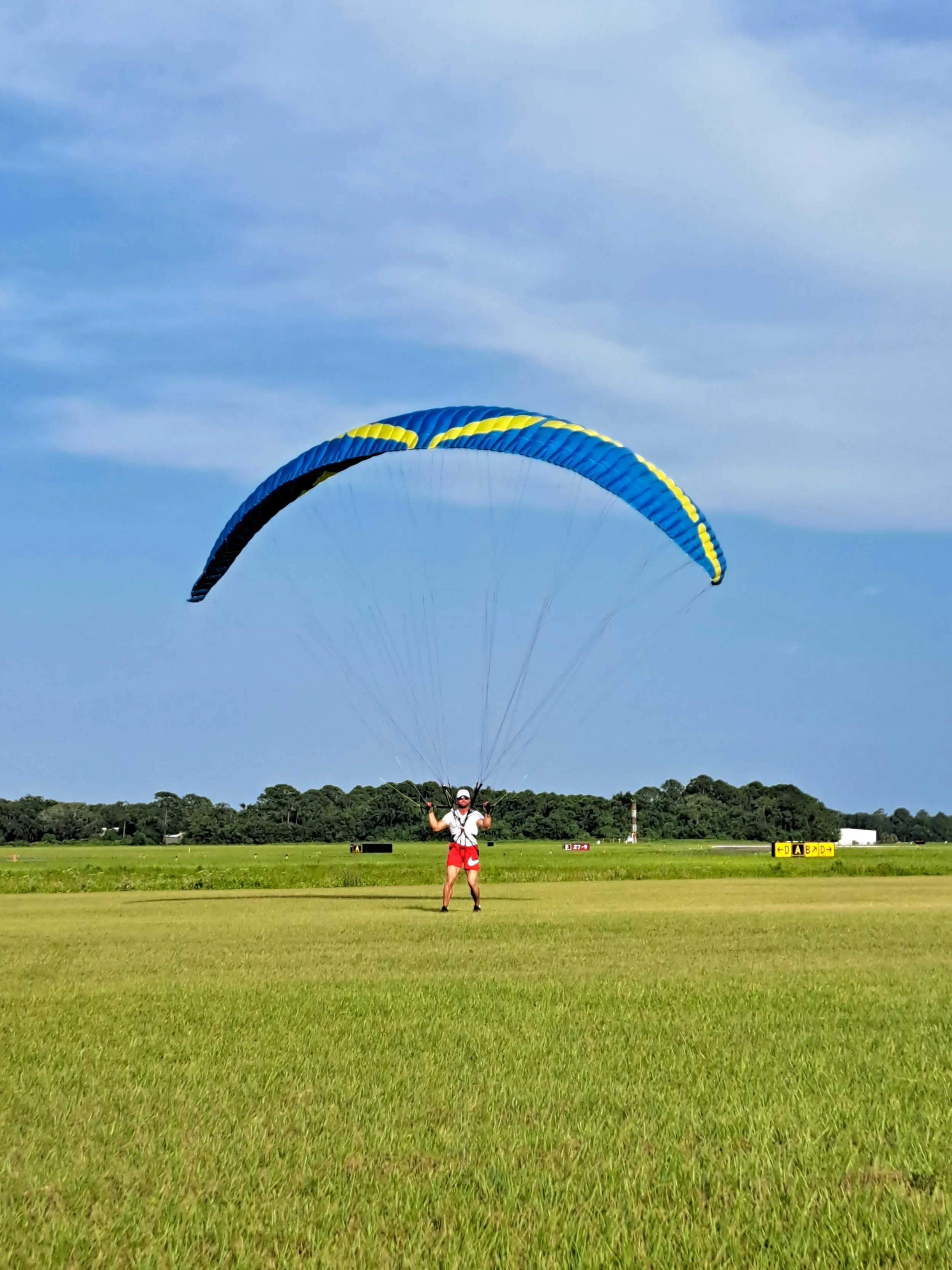 A person standing on a grassy field with a large parachute or canopy above them, preparing for paragliding or parachuting, with trees and a runway or airstrip in the background under a blue sky.
