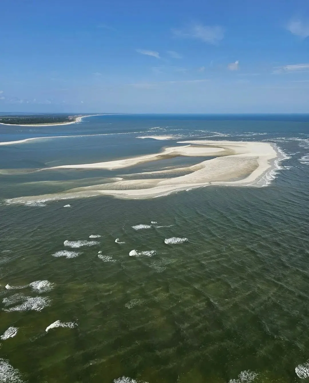 Aerial view of a coastal area with sandbars and tidal flats extending into the ocean under a partly cloudy sky.