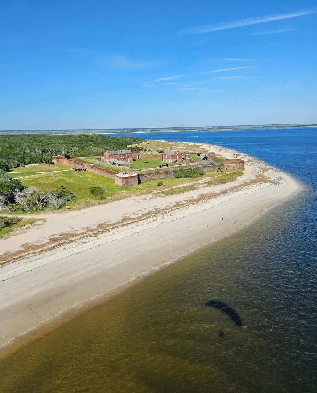 Aerial view of a historic coastal fort with brick walls and cannons, situated on a grassy area with a sandy beach and water nearby, under a clear blue sky.