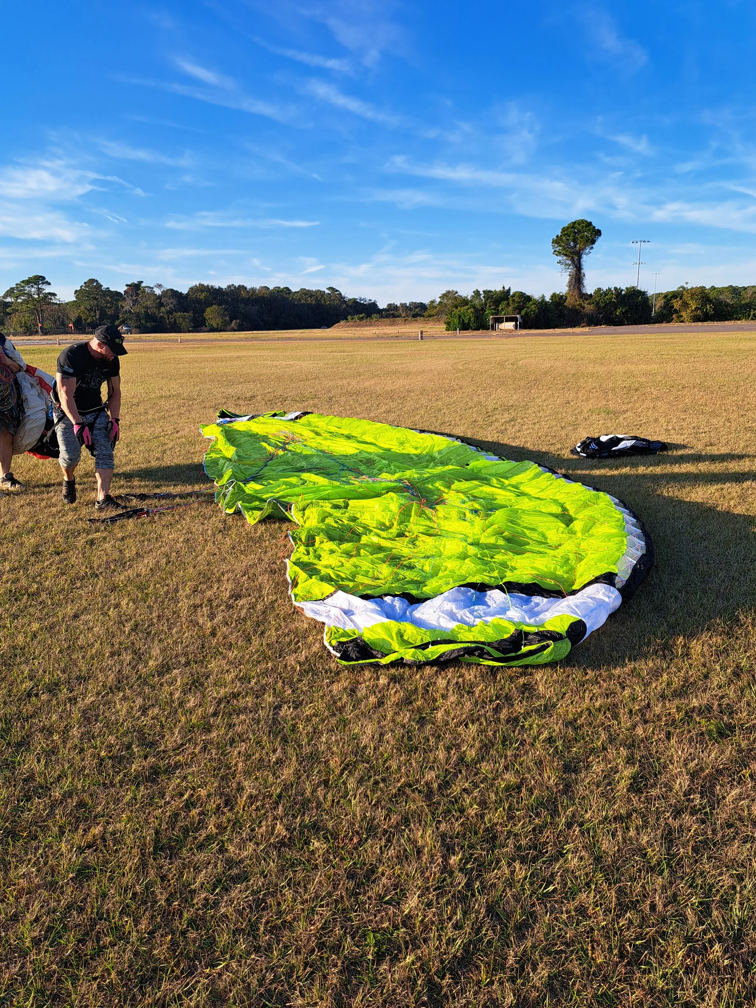 A person preparing a yellow and black paraglider on a grassy field with trees in the background under a blue sky.