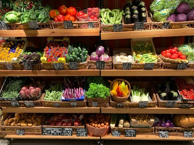 Display of fresh vegetables and fruits on store shelves. Top shelf has tomatoes, strawberries, bananas, zucchinis, and purple cabbages. Middle shelf includes grapes, cucumbers, onions, and tomatoes. The bottom shelf features potatoes, radishes, carrots, leeks, and more.