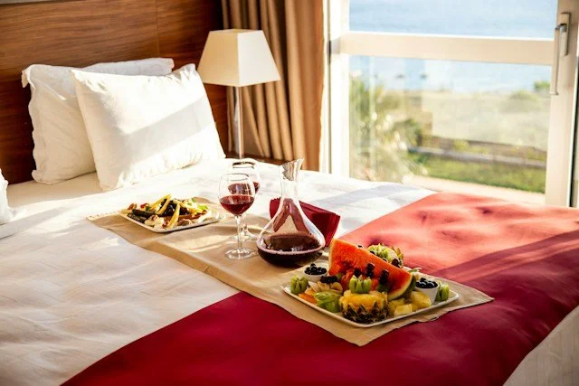 A hotel bed with a breakfast tray that includes a glass of red wine, a carafe of wine, a bowl of fruit, and a plate of food, near a large window with a view of the ocean and palm trees.