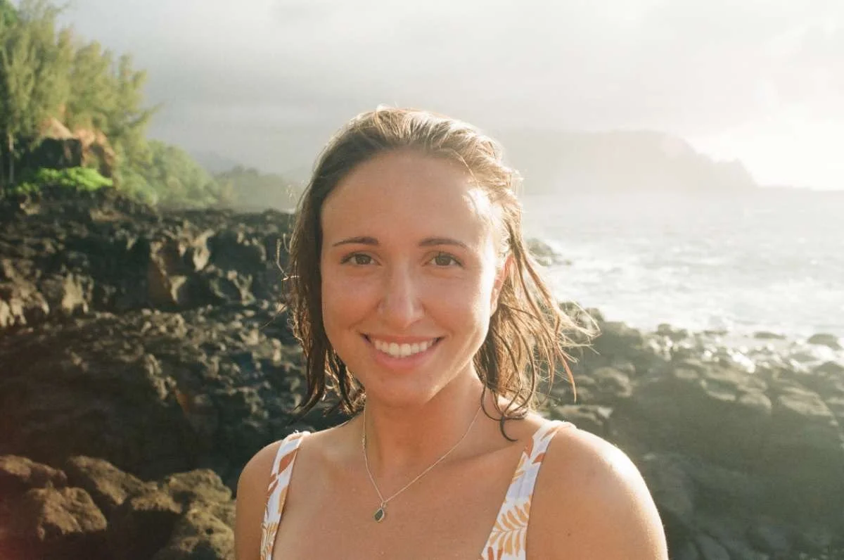 A young woman with wet hair smiling at the camera on a rocky beach with ocean waves and a cloudy sky in the background.