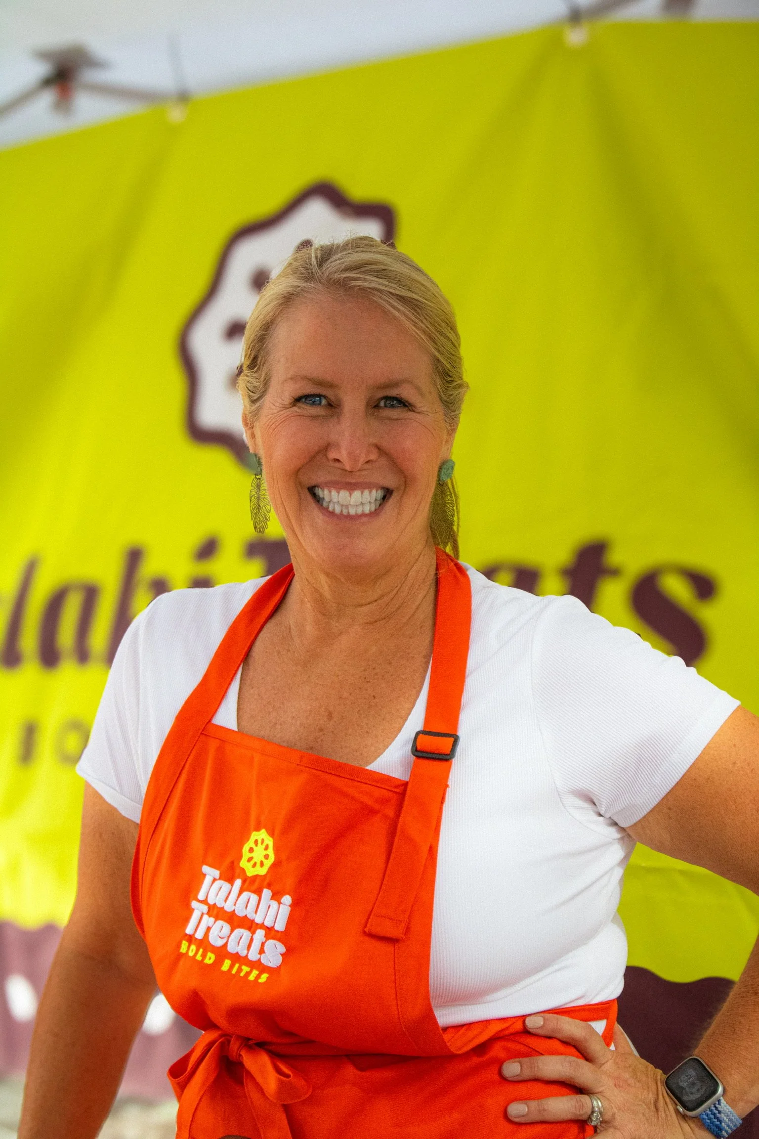 A woman smiling wearing a white shirt and orange apron with the logo and text 'Talahi Treats Bold Bites' in front of a chartreuse backdrop with blurred text.