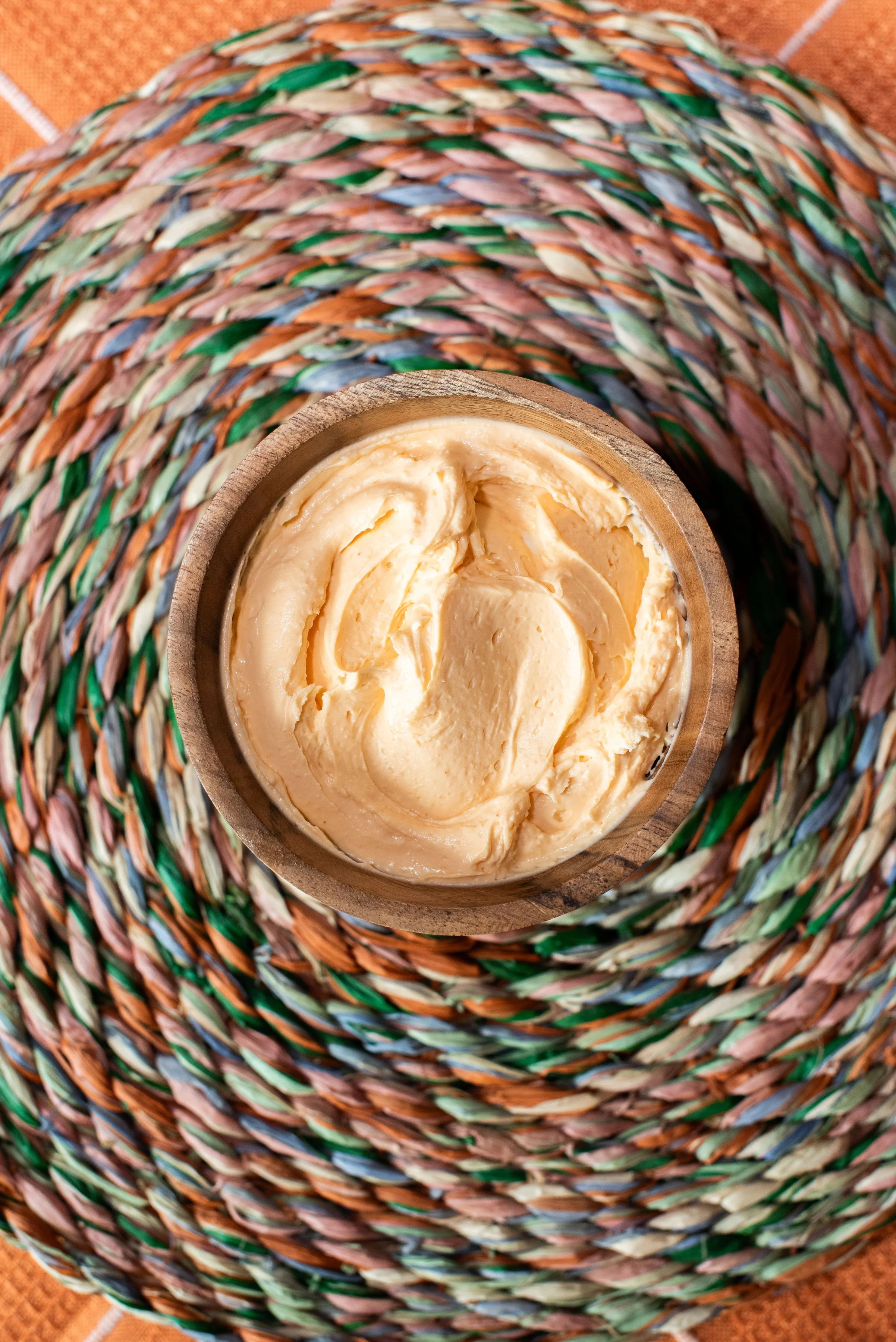 A wheel of soft, creamy cheese in a wooden bowl, placed on a woven, multicolored basket, with a woven orange textile underneath.