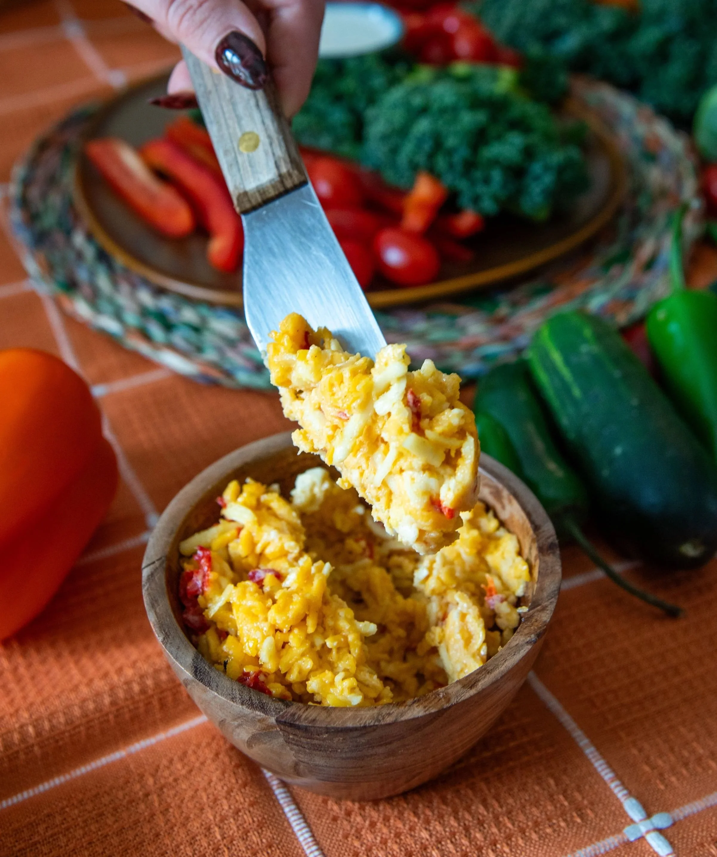 A spoonful of pimento cheese dip being held over a wooden bowl filled with scrambled eggs with red bell pepper pieces. In the background, there are fresh green broccoli, cherry tomatoes, and other vegetables on a brown table with an orange cloth.