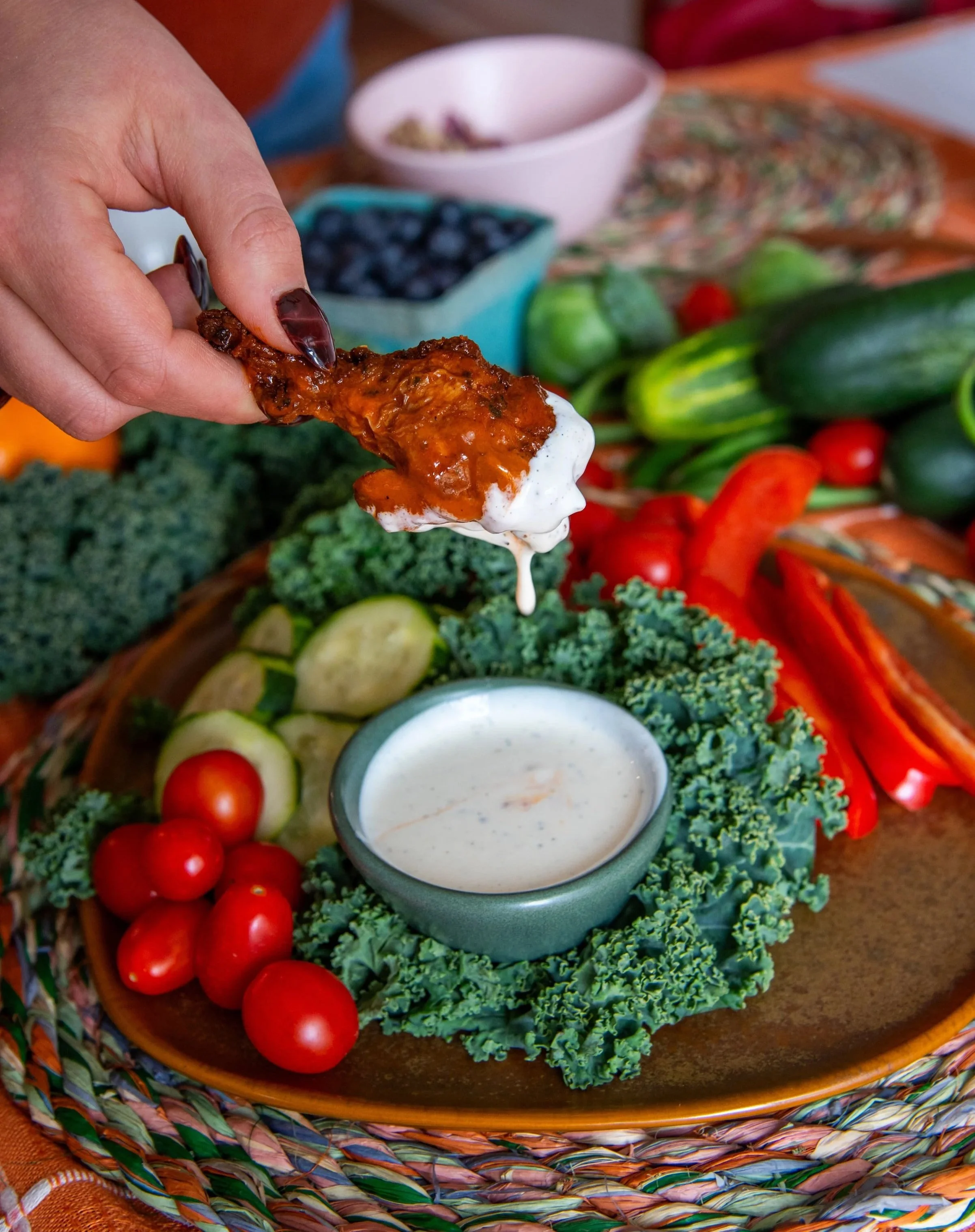 Fried chicken wing being dipped in a ranch dipping sauce with fresh vegetables and berries on a colorful woven tray.
