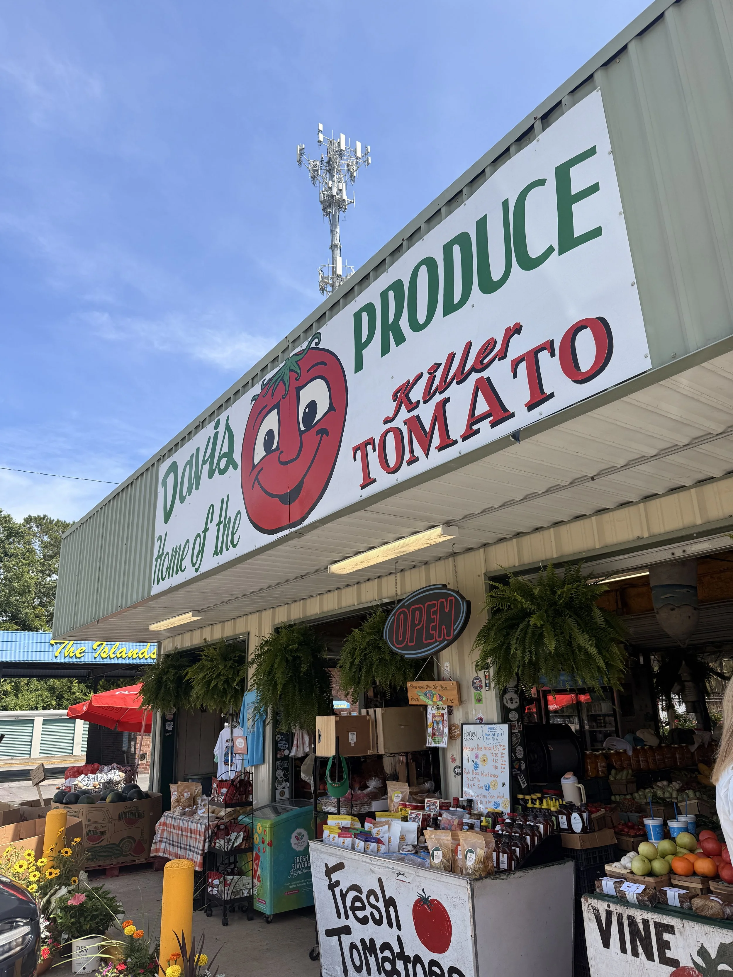 Farmers market stand selling fresh tomatoes, watermelons, and other produce, with signs advertising 'Fresh Tomato' and 'Vine' and an 'Open' sign, under a clear blue sky.