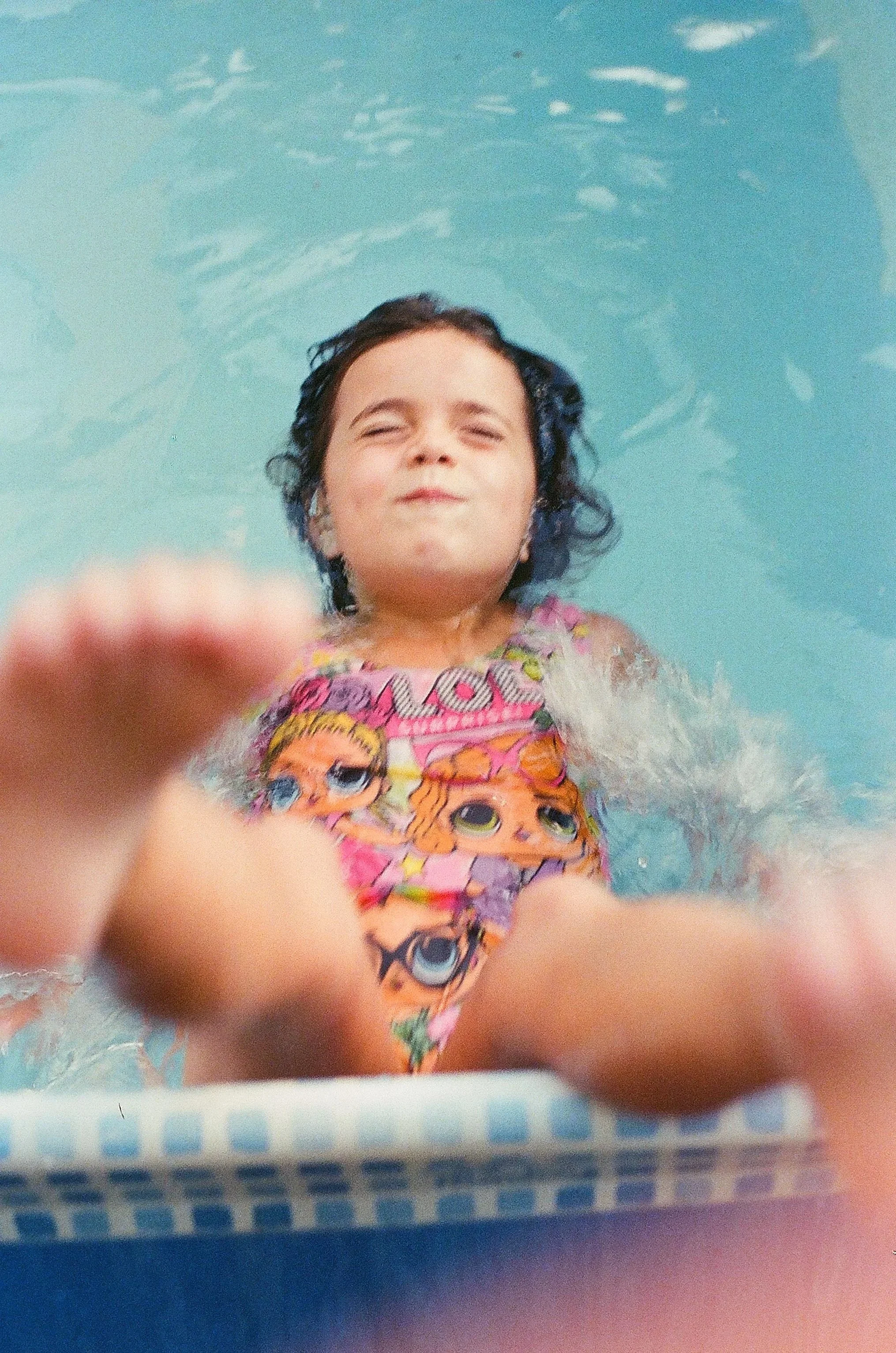 A young girl with dark hair and a colorful shirt with cartoon characters is floating on her back in a swimming pool, smiling with her eyes closed. Her face is relaxed and she appears to be enjoying herself.