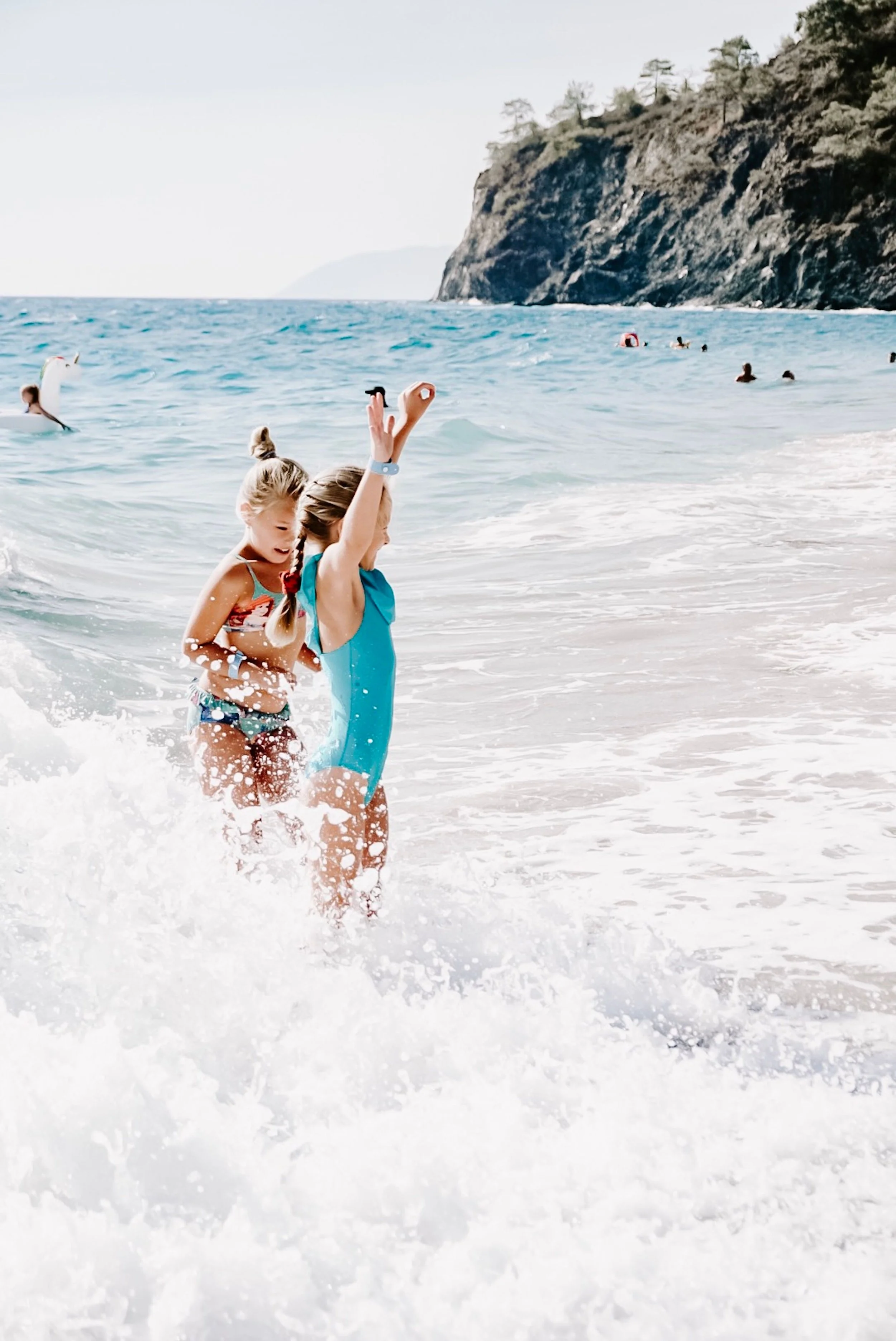 Two young girls playing and splashing in the waves at the beach, with a rocky cliff and some trees in the background.