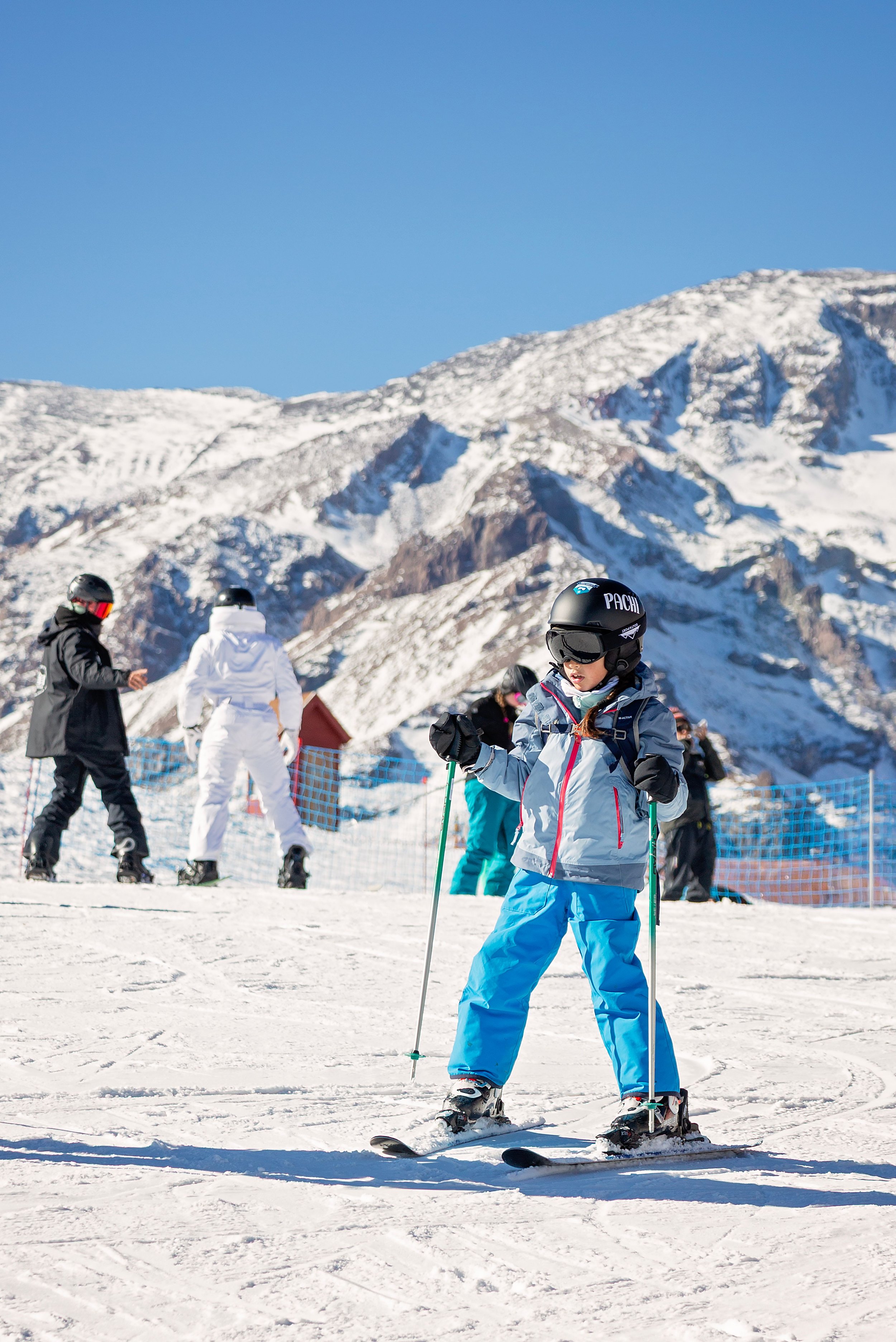 A young girl wearing a helmet and winter ski gear on skis at a snowy ski resort with snow-covered mountains in the background.