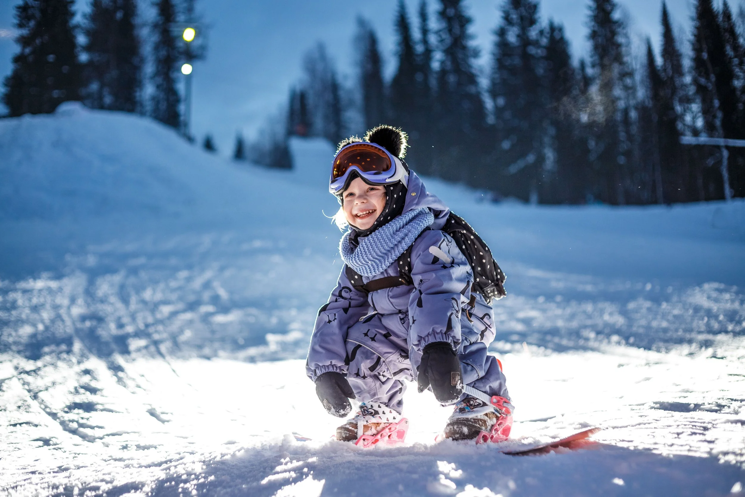 A young girl wearing a winter jacket, snow pants, gloves, a scarf, and ski goggles is snowboarding on a snowy slope during evening or late afternoon, with snow-covered trees and faint light in the background.
