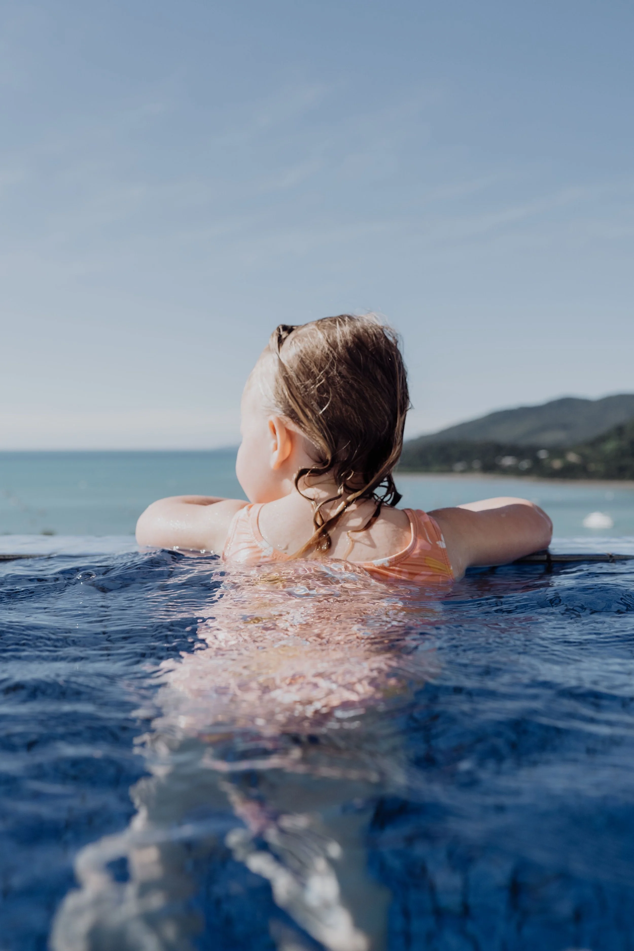 Person with wet hair resting arms on pool edge, overlooking ocean and mountains under blue sky.