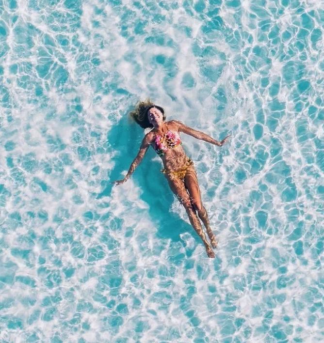 A woman lying in clear blue ocean water, smiling, wearing a colorful swimsuit and yellow shorts.