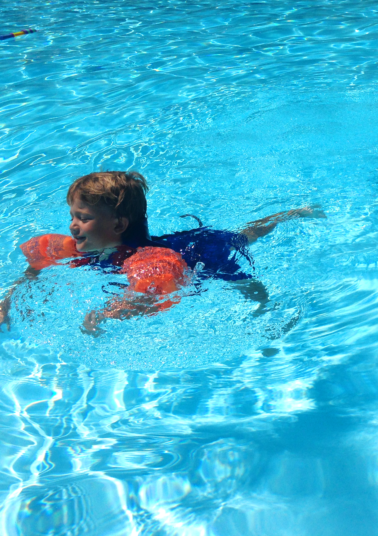 A young boy swimming in a bright blue pool, wearing orange floaties and a dark swimsuit, with sunlight creating reflections on the water.