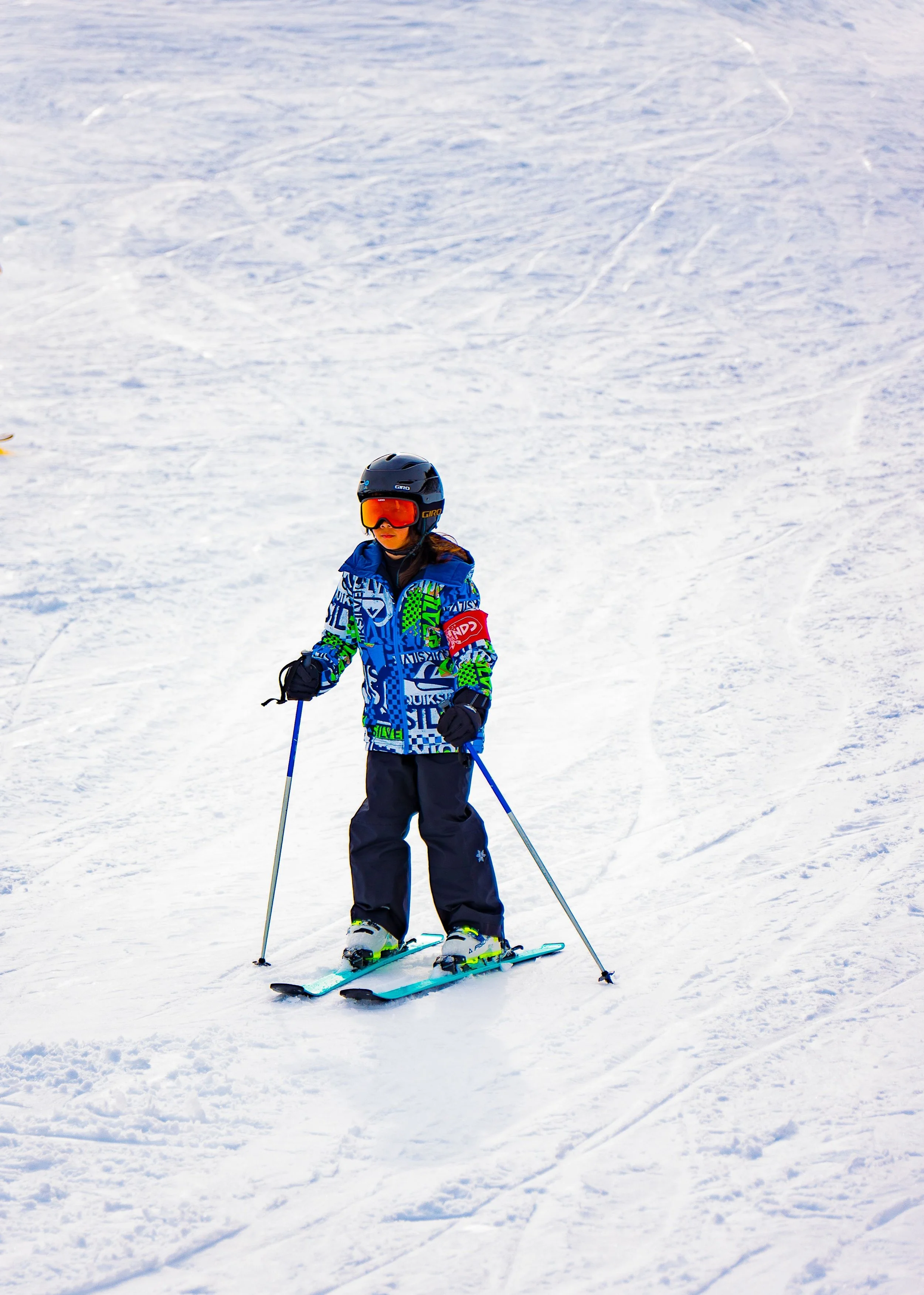 A young person standing on skis on snow, wearing a black helmet, orange ski goggles, a blue patterned ski jacket, dark ski pants, and gloves, holding ski poles, with ski tracks visible in the snow.