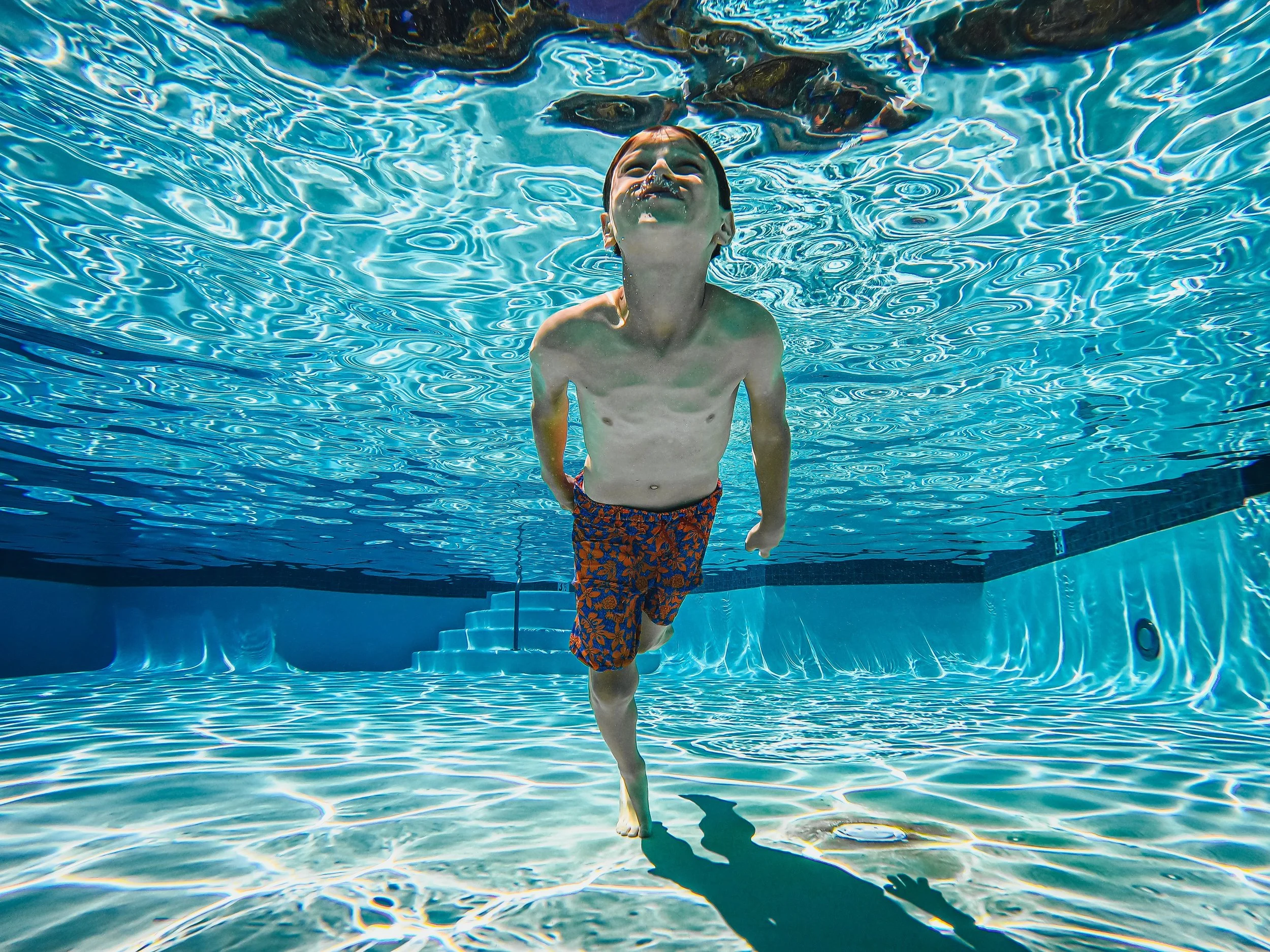 Child with wet hair and arm behind back, wearing swim trunks with a floral pattern, standing in a swimming pool underwater, smiling and looking up.