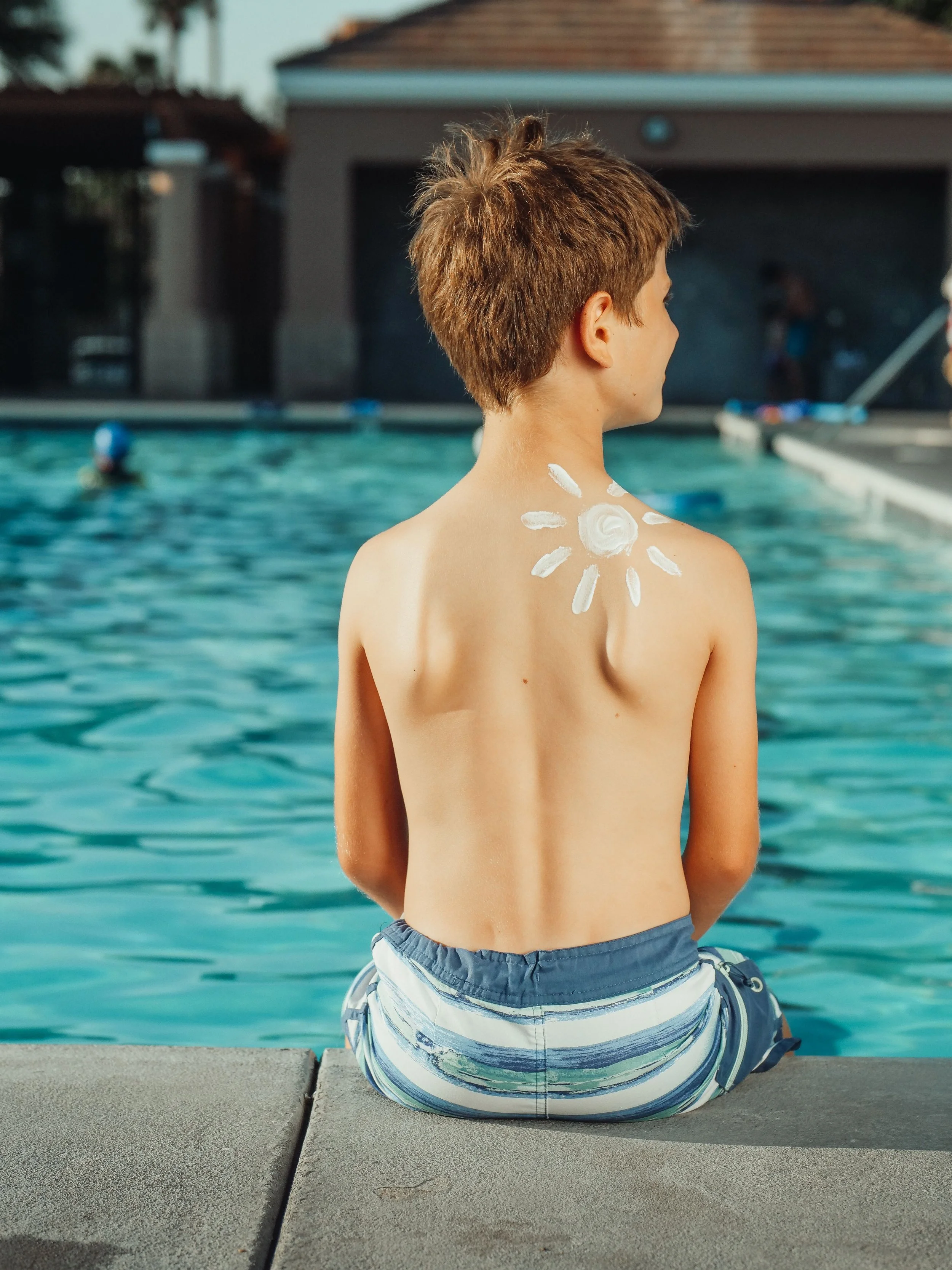 A boy with short brown hair, sitting on a concrete poolside edge, looking at the swimming pool. He has white paint or sunscreen painted on his back in the shape of a sun.