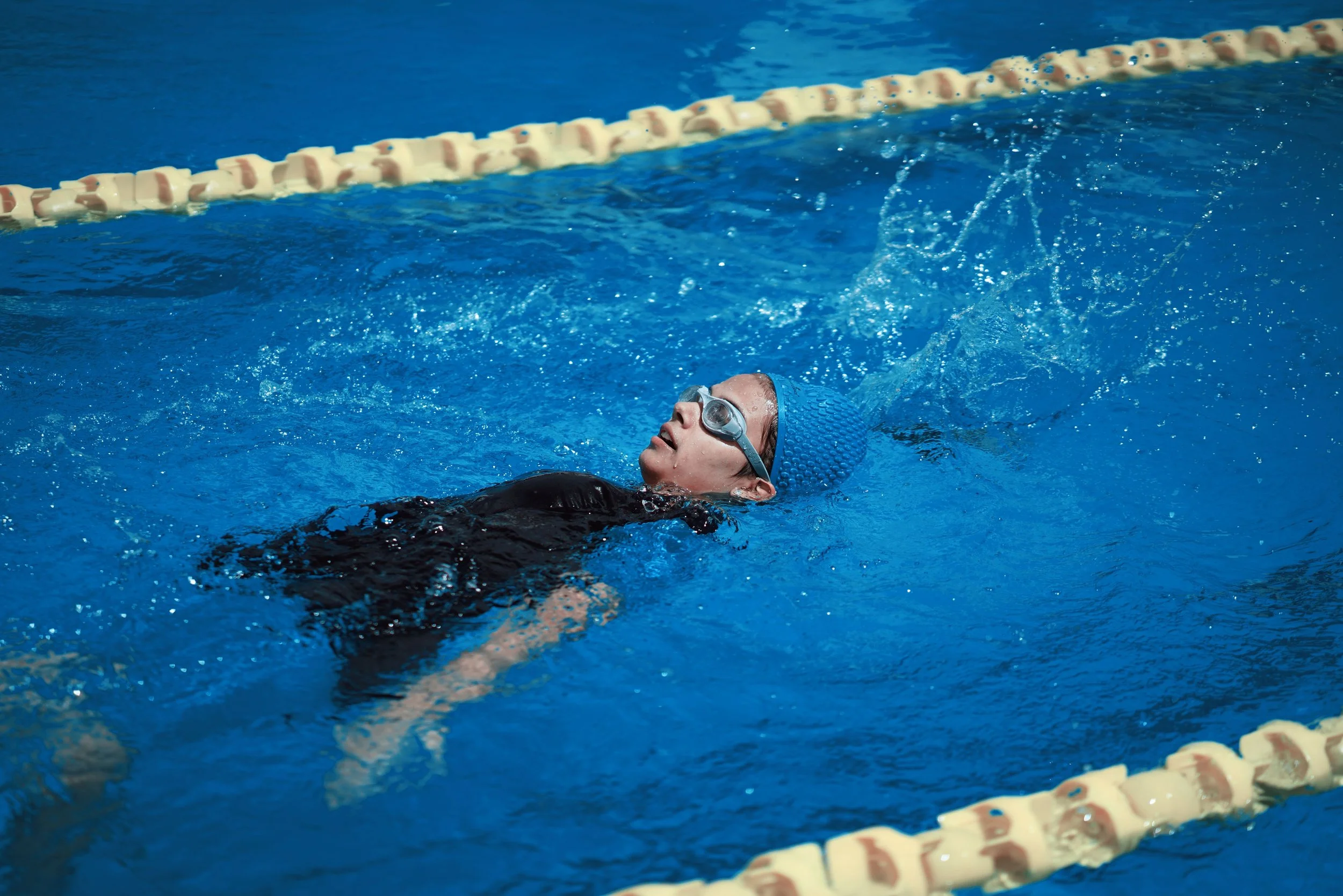 A swimmer wearing goggles and a blue swim cap is swimming in a pool near a lane divider.